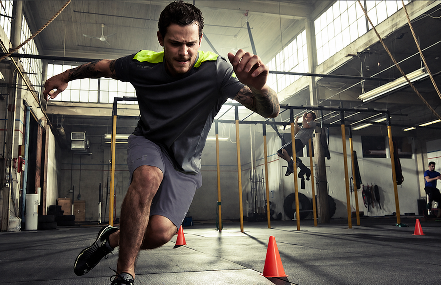 A man in athletic wear sprints through an agility course inside a gym, wearing a dark gray t-shirt with neon yellow-green shoulder panels and gray shorts. The background shows other gym equipment and another person training.