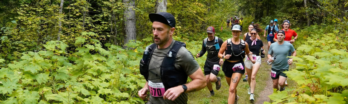 A group of trail runners racing along a forest path, with a man in the foreground wearing a black cap, gray t-shirt, and hydration vest with a race bib.