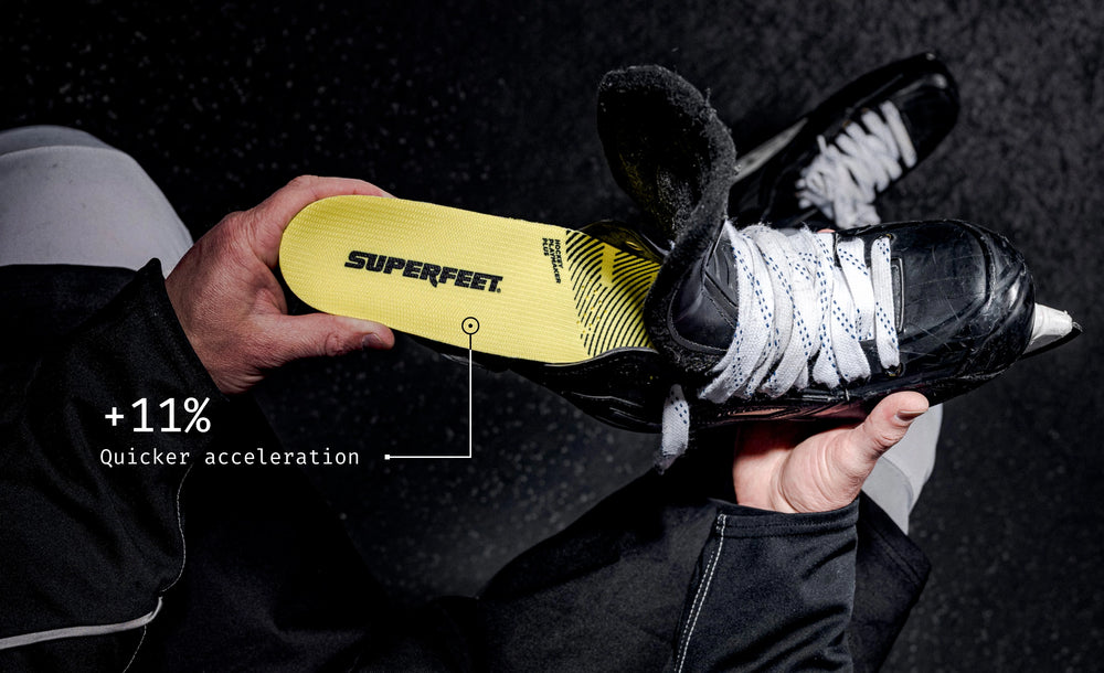 Person holding a black hockey skate with a yellow insole labeled 'Superfeet' on a dark background.