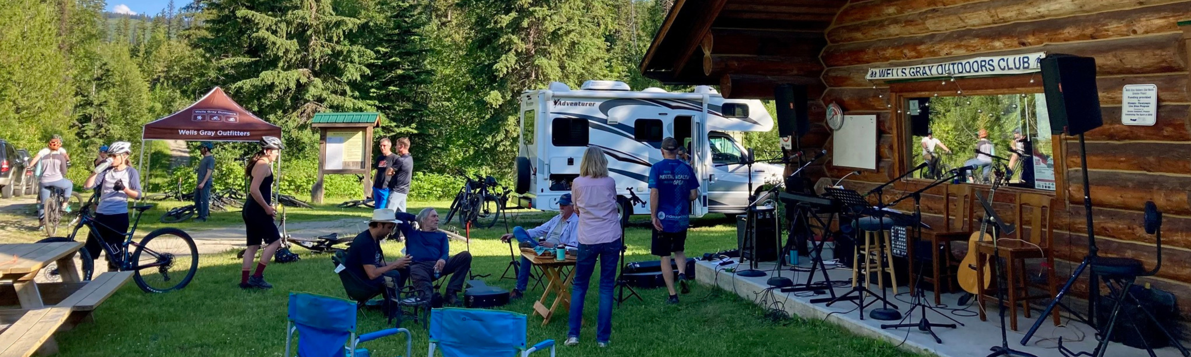 Outdoor gathering with people around a campsite, RV, and wooden cabin.