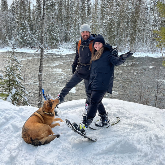 A young couple and their dog using rental snowshoes from Wells Gray Outfitters standing on snow covered ground in front of the Murtle River in Wells Gray Park.